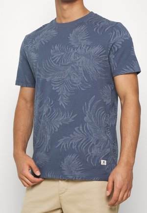 Mann trägt ein blaues T-Shirt mit kurzen Ärmeln und strukturierten tropischen Blattmustern sowie beige Hosen vor einem schlichten Hintergrund.