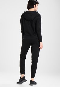 Crna hoodie uparena s odgovarajućim jogger hlačama, oboje izrađeno od mekanog materijala. Značajke uključuju rebraste manžete i kapuljaču sungee. Nosi se crne sportske cipele.