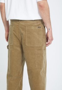 Pantalons en velours côtelé kaki avec plusieurs poches, tissu texturé et coupe décontractée. Présente une petite étiquette de marque à l'arrière.