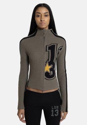 Nő bordázott cipzáras pulóverben, amelyen fekete és sárga csillaggrafika látható, valamint fekete nadrágban, amelyen a „LDN 13” felirat szerepel a combján.