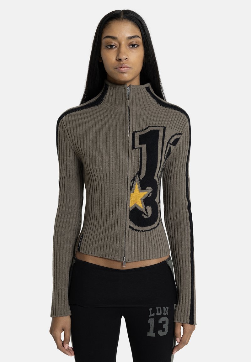 Γυναίκα που φοράει ριμπ πλεκτή ζακέτα με φερμουάρ, με μαύρο και κίτρινο γραφικό αστέρι, και μαύρο παντελόνι με το κείμενο "LDN 13" στο μηρό.