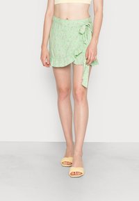 Saia envolvente floral em verde claro com bainha volátil e detalhe de atilho lateral, feita de tecido suave, usada com sandálias amarelas de bico quadrado.