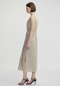 Abito senza maniche beige con una parte frontale testurizzata, spacchi laterali e vita regolabile. Indossato con tacchi neri con lacci. Design semplice, tessuto leggero.