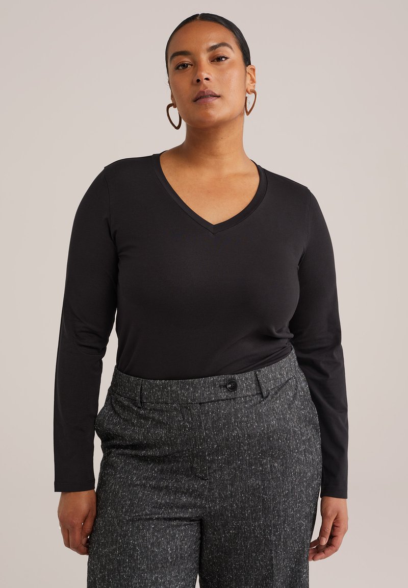 Mujer vestida con una camiseta negra de manga larga y cuello en V y pantalones grises texturizados, de pie contra un fondo claro y liso, de frente.