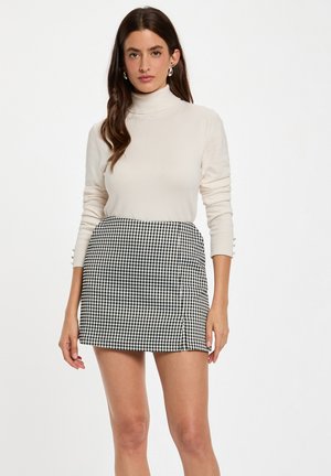 Maglione a collo alto color beige chiaro abbinato a una mini gonna a quadretti neri e bianchi. La gonna presenta una apertura laterale e dettagli con bottoni sulle maniche.