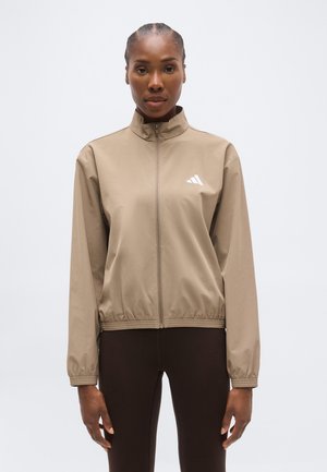 Femme portant une veste beige zippée avec poignets et taille élastiques, debout les bras détendus devant un fond clair uni.