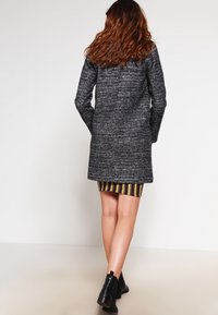 Lungo cappotto grigio scuro con motivo strutturato, caratterizzato da una linea dritta. Indossato sopra un abito a righe di lunghezza al ginocchio e stivaletti neri.