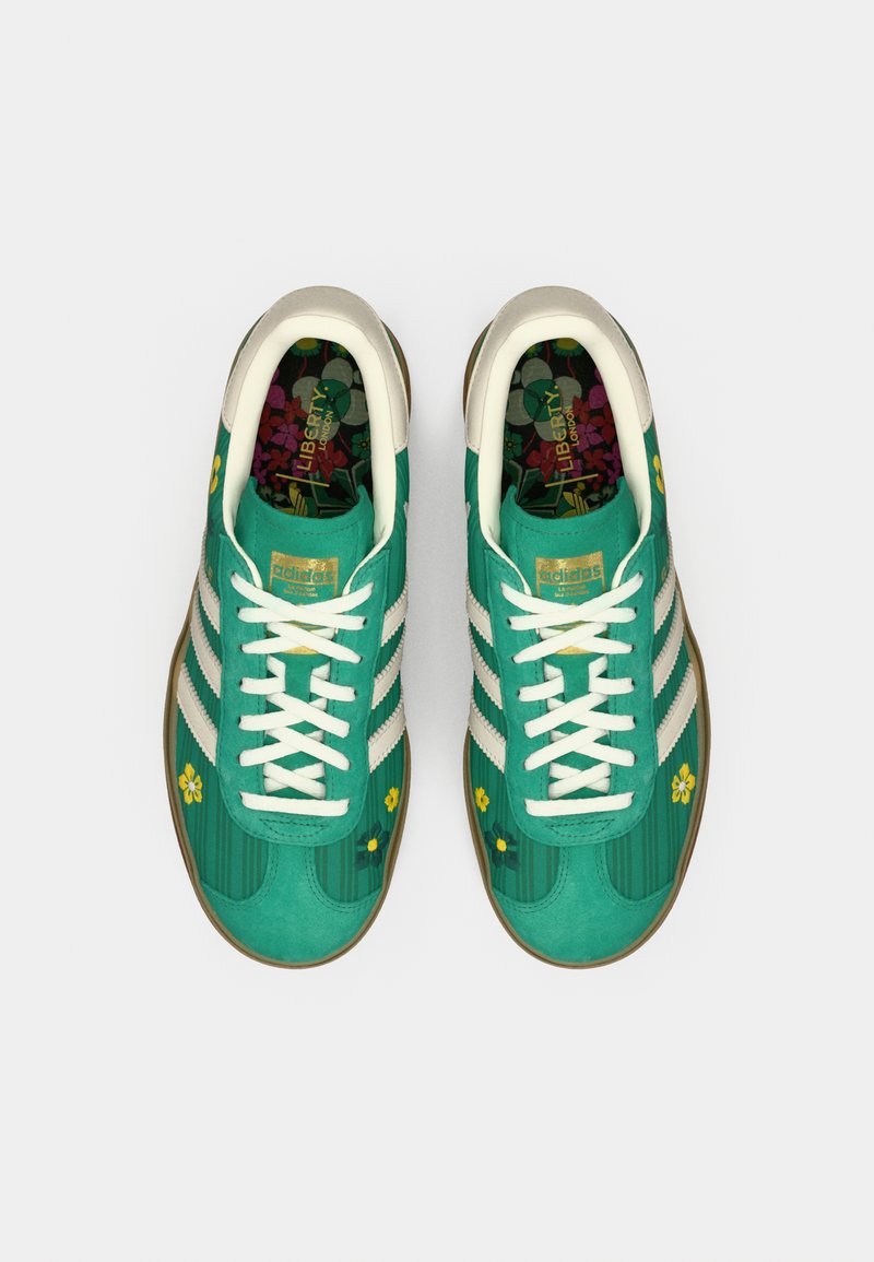 Zapatillas verdes con textura de ante, franjas blancas, detalles florales y una plantilla con estampado. Cuentan con una suela de goma beige y cordones.