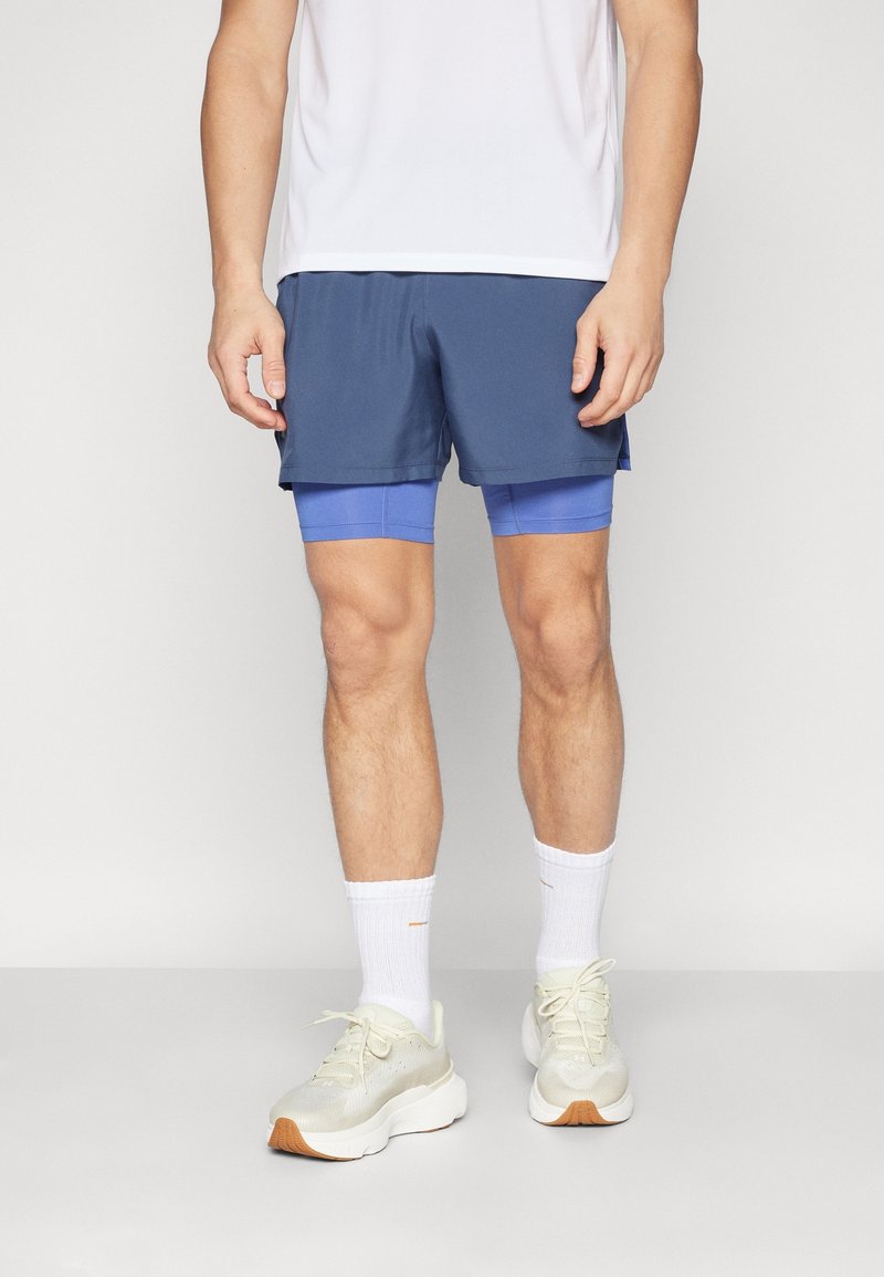 Homme portant un short de sport bleu superposé, des chaussettes blanches et des chaussures de course beige, debout devant un fond uni.