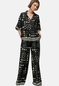 Schwarzes Seidenpyjama-Set mit großen weißen und goldenen Punkten. Das Oberteil hat einen V-Ausschnitt und kurze Ärmel; die Hose ist weit geschnitten und hat gestreifte Akzente.