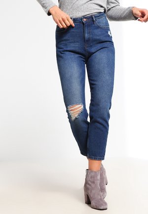 Frau trägt hoch taillierte blaue Jeans mit einem zerrissenen Knie, ein graues Langarmshirt und graue Veloursleder-Stiefeletten, steht vor einem weißen Hintergrund.