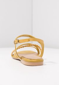 Sandales en cuir jaune avec des fines brides, des détails tressés et une fermeture à boucle. Elles disposent d'une semelle plate beige et d'une finition lisse.