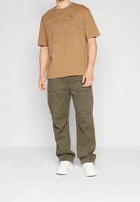 Brun bomulls-t-shirt med en grå krokodilgrafik, tillsammans med olivgröna cargobyxor som har flera fickor och vita sneakers.