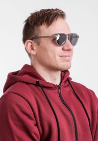 Šedé sluneční brýle s kulatými rámy, černými skly a texturovanými detaily na stranách. Model má na sobě vínovou mikinu na zip s černými šňůrkami.