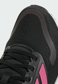 Black athletic shoe featuring a textured mesh upper, black laces, and a pink accent on the side. Smooth rubber sole and reinforced heel.