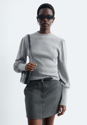 Femme portant des lunettes de soleil noires, un pull gris à manches longues, une jupe gris foncé, une ceinture noire et un sac à épaule noir sur un fond uni.