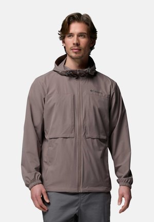 Junger Mann, der eine taupefarbene Kapuzenjacke mit Brusttaschen vorne und elastischen Bündchen trägt, steht vor einem schlichten hellen Hintergrund.