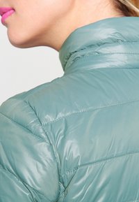 Närbild av en persons axel och nacke som bär en ljusgrön vadderad jacka med hög krage och ljusrosa läppstift synligt.