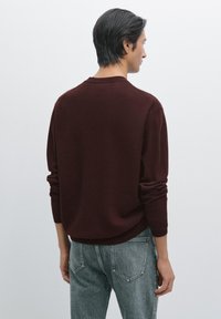 Maglione di lana bordeaux con maniche lunghe e polsini arrotolati, abbinato a jeans di denim grigio chiaro, visto da dietro.