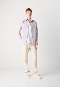 Helle lila Hemd mit Knöpfen über einem weißen T-Shirt, kombiniert mit beigen Hosen und hellen Sneakers. Glattes Material, lässige Passform und lockerer Look.