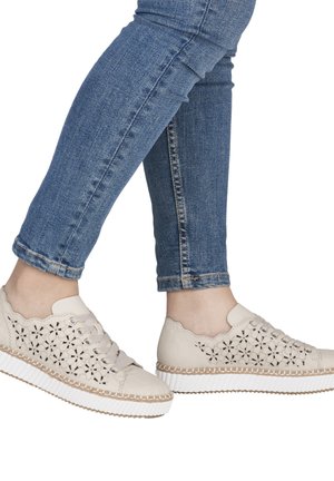 Feet wearing white lace-up shoes with cut-out floral patterns paired with blue denim jeans, shown against a white background.