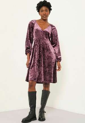 Plüschiges, mittellanges, burgunderrotes Samtkleid mit V-Ausschnitt, langen Ärmeln und ausgestellt Rock; kombiniert mit kniehohen schwarzen Lederstiefeln.