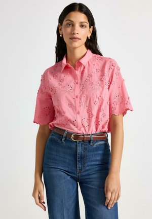 Lyserød kortærmet skjorte med broderi og blomstermotiver, krave og bølgede kanter, kombineret med blå højhøjede denimjeans.