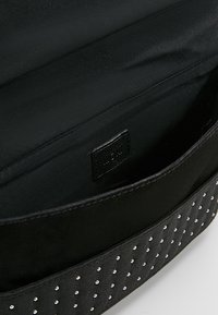 Černá kabelka s hladkým povrchem a texturovaným pruhem s stříbrnými cvočky. Interiér vyložený černou tkaninou; logo viditelné.