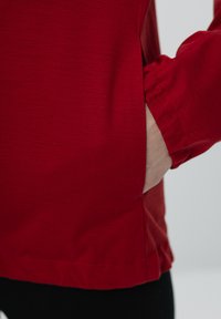 Czerwona bluzka z długim rękawem o gładkiej fakturze, z bocznym rozcięciem blisko dołu i mankietem. Ręka umieszczona w kieszeni.