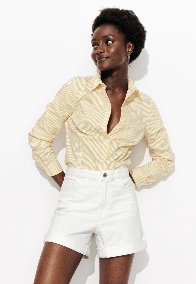 Chemise à boutons rayée jaune et blanche, portée avec un short blanc à ourlet roulé, présentant un col et des manches longues.