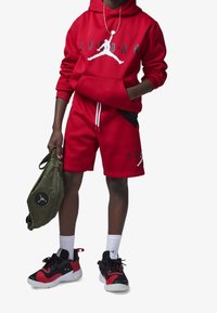 Sudadera con capucha roja y pantalones cortos con detalles y logo en negro; zapatillas negras y rojas; bolso verde oliva con logo. Calcetines blancos.