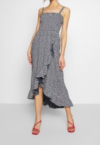 Kvinna i en marinblå blommig midiklänning med volangkant och röda spetsskor med hög klack, stående mot en enfärgad ljus bakgrund.