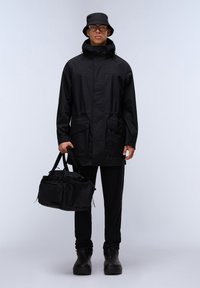 Svart vattentät jacka med justerbar huva, två framfickor, tillsammans med en matchande bucket hat och svart duffelväska, bärs över svarta byxor och stövlar.