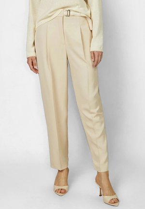 Frau trägt beige plissierte maßgeschneiderte Hose mit passendem Gürtel und cremefarbene offene High-Heel-Mules, stehend vor einem schlichten Hintergrund.