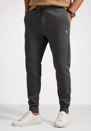 Homme portant un pantalon de jogging gris foncé avec cordon et un logo blanc sur la cuisse, des baskets blanches, un pull beige et une montre-bracelet au poignet gauche.