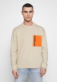 Beige hosszú ujjú póló, bal mellen narancssárga cipzáras zsebből, pamutból készült, kerek nyakkivágással és laza fazonnal.