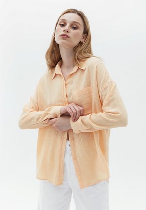 Knopenhemd in licht perzik, gemaakt van een gestructureerde stof. Het heeft een borstzak en lange mouwen, gedragen over witte broek.
