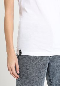 Biała bawełniana koszulka z okrągłym dekoltem, wyeksponowana obok strukturalnych, szarych dżinsów. Posiada czarną metkę marki umieszczoną w lewym dolnym rogu.