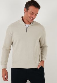 Heled beež värviline terry-kangast tõmblukuga pullover, ribakujuline tekstuur, kõrgem kaelus ja pikad varrukad, kantud valge T-särgi peal.