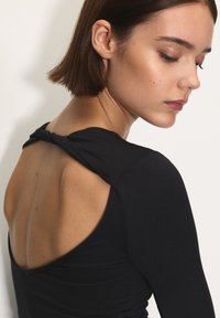 Rövid barna hajú nő fekete, csomózott, nyitott hátú felsőben, lefelé és kissé oldalra nézve, fehér háttér előtt.