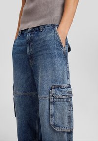 Cargojeans i medelblå denim med stora sidofickor, lös passform, synlig sömsömn och lätt blekning över hela tyget.