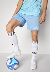 Himmelblaue Sportshorts mit gelben Akzenten und weißem Logo, kombiniert mit weißen Fußballschuhen und knielangen weißen Socken mit schwarzen Streifen, auf einem gemusterten Fußball.