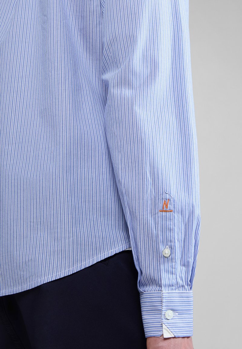 Koszula w paski w jasnoniebieskim i białym kolorze, z zapięciem na guzik przy mankietach oraz haftowanym logo na rękawie. Gładka tekstura materiału.