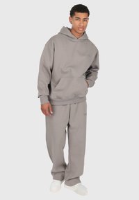 Giovane uomo che indossa un ampio hoodie grigio e pantaloni della tuta coordinati, con sneakers bianche, in piedi su uno sfondo semplice.