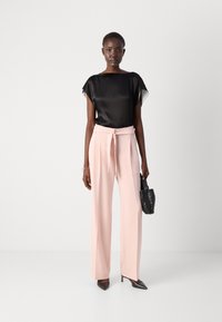 Blusa nera in satin con maniche corte, abbinata a pantaloni a vita alta e larghi di un delicato rosa pastello. Il modello tiene una piccola borsa nera con texture.