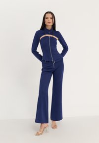 Navyblauw getailleerd langemouwen top met rits aan de voorkant en uitsnijdetail, gecombineerd met bijpassende wijde broek. Doorzichtige sandalen met hak.