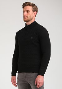 Schwarzer Pullover mit einem Viertel-Reißverschluss-Kragen, strukturiertem Rautenmuster auf der Vorderseite sowie geripptem Saum und Bündchen.
