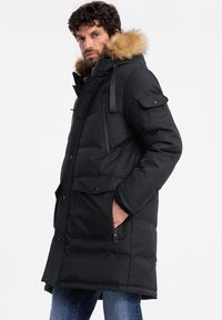 Parka nera imbottita con cappuccio rifinito in pelliccia, multiple tasche, chiusure con zip e bottoni, e cinturini regolabili. Texture esterna liscia.