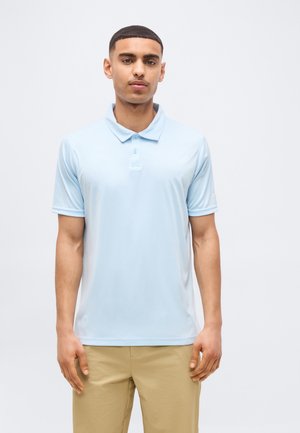 Junger Mann trägt ein hellblaues Poloshirt und beige Hose, steht vor einem schlichten weißen Hintergrund.