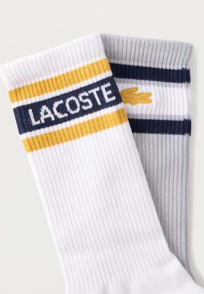 Deux paires de chaussettes côtelées hauteur cheville, une blanche à rayures jaunes et bleu marine avec le texte "LACOSTE", une grise à rayures bleu marine et blanches avec un logo crocodile jaune.